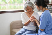 A young female doctor in blue scrubs holds hands with an older female patient, both sitting on a couch. (iStock/BongkarnThanyakij)